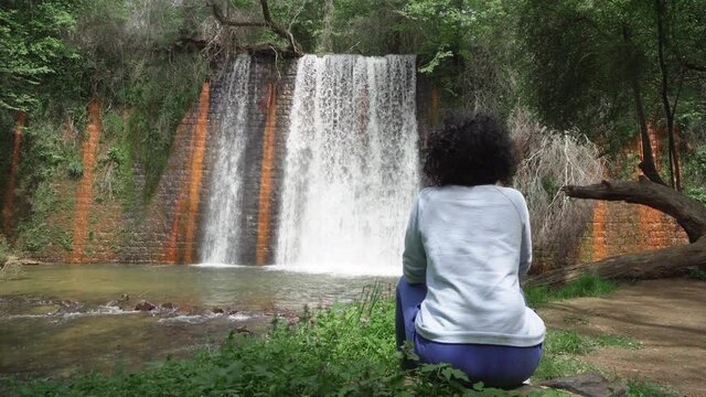 Young woman with curly hair sitting on the mossy rocks and enjoying the view of the jungle waterfall and creek. Travel and freedom concept