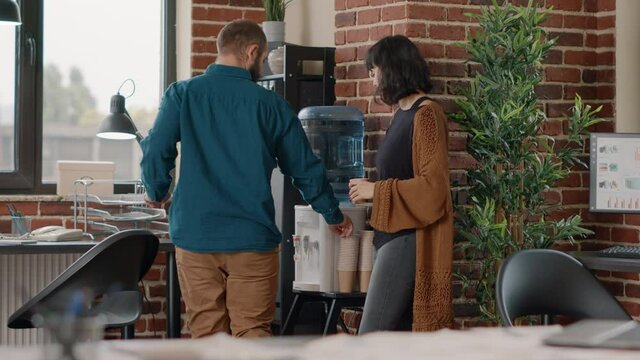 Colleagues meeting on break while having refreshment from water cooler in startup office. Man and woman having discussion while drinking fresh water for hydration from dispenser.
