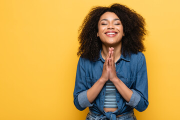 positive african american woman with closed eyes showing please gesture isolated on yellow