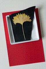 autumn ginkgo leaf on black leather inside a shallow white dish and arranged on a red and white paper background