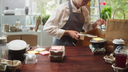 Food delivery service. Woman packaging meals, putting lunch boxes with stickers into paper bag. Preparing take away containers. Healthy eating habits. Online contactless food shopping.