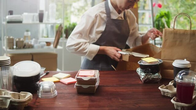 Food delivery service. Woman packaging meals, putting lunch boxes with stickers into paper bag. Preparing take away containers. Healthy eating habits. Online contactless food shopping.