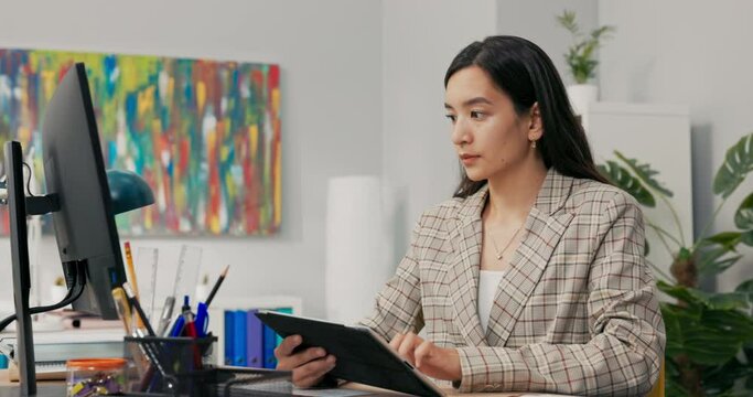 A conscientious organized office worker in a large corporation sits at desk synchronizing documents on tablet with computer, comparing data, clicking the screen, scrolling, focused on her work