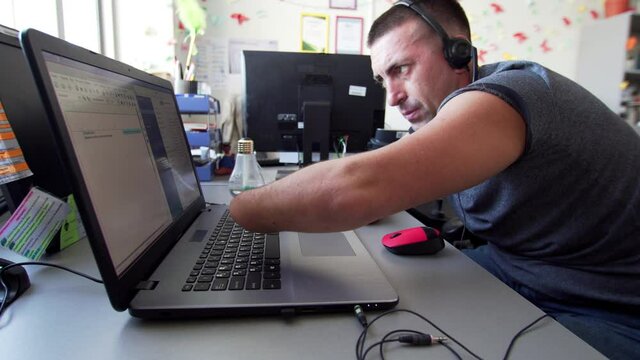 Portrait of a caucasian man in headphones with microphone with amputated two stump hands and legs types at the laptop. A man with a disabiity in wheelchair works in call-center at the laptop