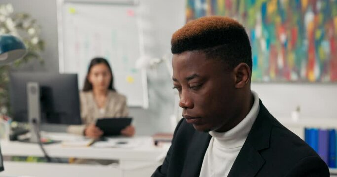 Dark-skinned man in jacket with colored hair works in company office fills out documents on compute no discrimination in background woman is answering email on tablet, everyday work in company