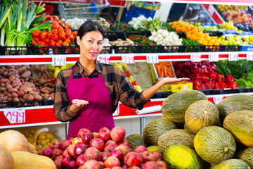 Polite friendly Asian saleswoman meeting customers in greengrocery store, offering fresh fruits and vegetables for sale