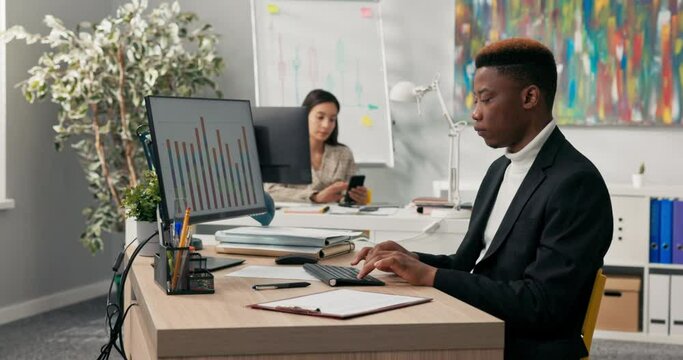 A focused dark-skinned man in suit spends time at work in front of office computer, completing charts on monitor, transcribing data onto pieces of paper, signing documents, completing applications