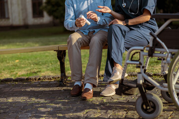 Obraz premium Cropped shot of elderly recovering male patient having conversation with a nurse, sitting together on the bench near rehabilitation clinic