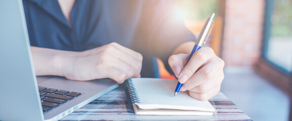 Business woman hand writing on a notepad with a pen and use a laptop computer in the office.