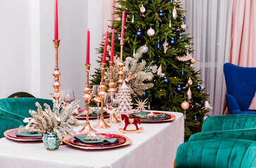 Luxurious New Year table setting. The room contains a Christmas tree, corduroy armchairs and a large table. On the table are golden plates, candles, and New Year decor