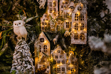 Fototapeta premium The toy owl sits on the background of toy wooden houses decorated with glowing garlands. New Year' decoration