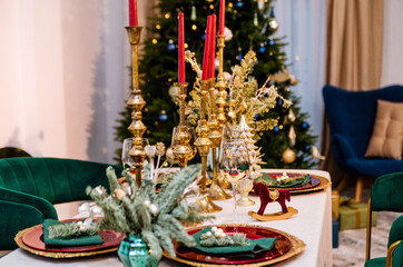 Luxurious New Year table setting. The room contains a Christmas tree, corduroy armchairs and a large table. On the table are golden plates, candles, and New Year decor