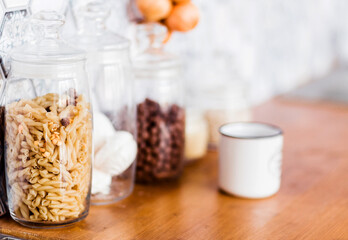 On the kitchen table, there are jars with various products - pasta, marshmallows, nuts. A jar of pasta is in focus of the camera.