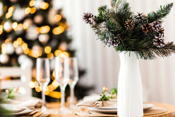 A white vase with Christmas tree branches stands on a festive New Year table against the background of a Christmas tree with a garland
