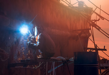 Side view of welder in protective workwear on scaffold is welding the old rusty vessel hull on overtime in shipyard area at sunset time