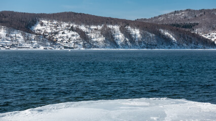 Ice-free river in winter. Ripples on the blue water. There is snow on the coastal hills. Ice on the shore. A sunny day. Siberia. Angara 