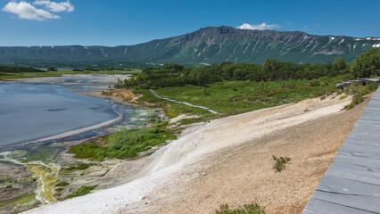 There is a blue thermal lake in the caldera of an extinct volcano.  Wooden paths for tourists are laid on the ground. A mountain range against the sky. Kamchatka. Uzon