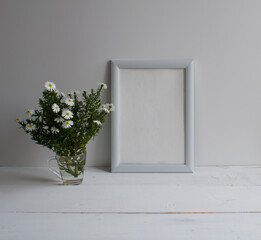 A mockup of a white empty frame, a bouquet of flowers on a wooden table against a light wall background.