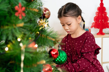 selective focus of an asian little girl preparing for xmas by a Christmas tree, putting decorations with concentration in the living room at home