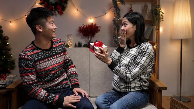 surprised asian wife receiving christmas present from hasband is smiling and covering mouth as she see inside the box on couch at a festive home interior