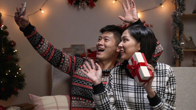 cheerful asian couple holding a Christmas gift while they are waving hi and making video call to their friends on mobile phone during Christmas holiday at home