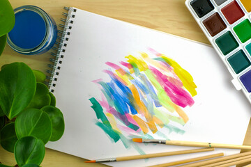 watercolor paints and brushes on an album sheet with paint samples next to a greenhouse plant