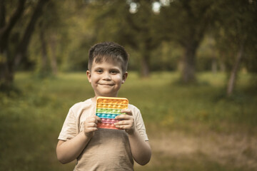 Smiling Boy With Rainbow Pop It Walks in the Park. Nice Kid Plays Pop It Outdoor. High quality photo