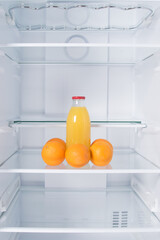 against the background of a white refrigerator, there is a bottle of juice and oranges on a glass shelf © kurgu128