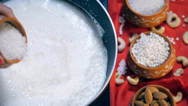 An Indian cook adding sugar into the milk mixed with white tapioca pearls. Closeup shot of sugar and Sabudana in earthen pots