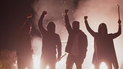 Four silhouettes of rebels in black masks with various edged weapons and with raised fists stand background of burning car, front view. Anarchists set fire to car in protest. Concept of protest