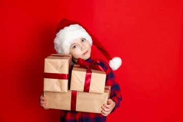 Happy holiday and Merry Christmas. Portrait of a boy in a cap with gifts in his hands on a red background. A place for text.