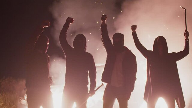 Four silhouettes of rebels in black masks with various edged weapons and with raised fists stand background of burning car, front view. Anarchists set fire to car in protest. Concept of protest