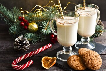 New Year or Christmas Eggnog cocktail - hot winter drink with milk with cinnamon, napkin and sweet candy in a glass mugs and fir tree branches with festive decorations, garland on wooden table