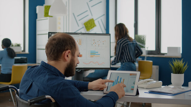 Paralyzed manager, businessman with disabilites handicapped immobilized man using computer and tablet in same time working in business start-up office sitting in wheelchair. Busy multitasking employee