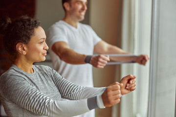 Cheerful sporty couple is training arms before window together at home and using resistance band