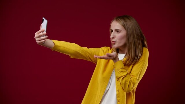 the girl takes a selfie on the phone, she really likes to take pictures, sends an air kiss, the background is red, she is wearing a white T-shirt and a yellow shirt, the background is red