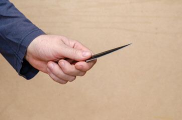 A hand with a knife against a brown background. Adult male holding a small double-edged knife.