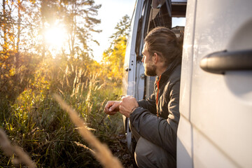 Man enjoying a morning coffee in the doorway of his camper van in autumn - obrazy, fototapety, plakaty