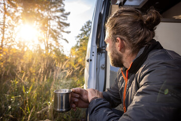 Man having a morning coffee in the doorway of his camper van in autumn