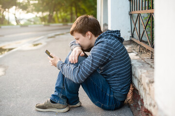 A lonely and sad teenage student, hiding in a closed pose from problems on the street in an urban environment with a mobile phone in his hands. The concept of social problems of youth.