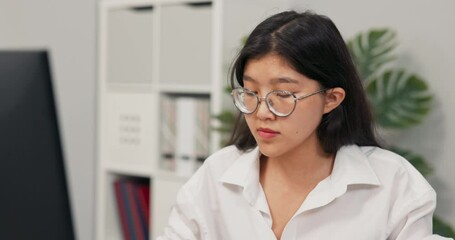 Beautiful secretary in glasses with characteristic Korean Asian beauty dressed in white shirt, sits in the office in front of the computer, working, in the background co-workers walk around company