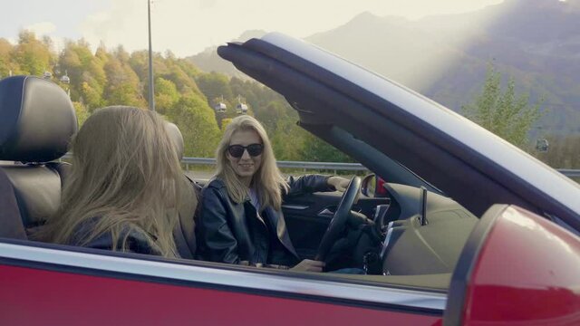 Two Caucasian girls are sitting and talking in a red convertible. Happy young girls in sunglasses are resting in the mountains.