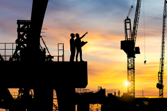 Silhouette of Engineer and worker checking project at heavy infrastructure building site background, construction site at sunset in evening time.