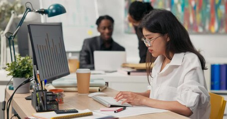 Beautiful girl with glasses with distinctive Asian beauty dressed in white shirt, sits in office in front of computer, working, in background co-workers are discussing, talking, afternoon at company