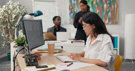Beautiful secretary in glasses with characteristic Korean Asian beauty dressed in white shirt, sits in the office in front of the computer, working, in the background co-workers walk around company