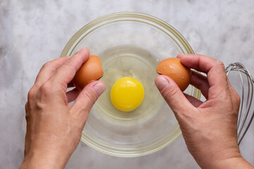 Top view of woman hand cracking raw brown egg into glass bowl on marble surface