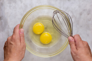 Top view of woman hands holding glass bowl with two raw yolks and whites in it and whisk on marble surface