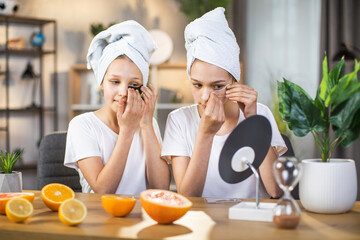 Two caucasian sisters wearing towel on head and white t-shirt sitting together at desk and applying patches under eyes. Beauty procedures for keeping youth.