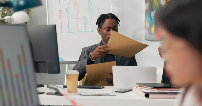 Busy dark-skinned boss in suit sits behind desk at computer, assistant brings him envelopes, letters, man opens, looks through correspondence, gives back to woman, leaves on desk