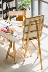Top view shot of drawing painting artist hobby workshop studio equipment on wooden table including canvas easel stand watercolor paintbrush pencils palette tray paper under strong light from outside